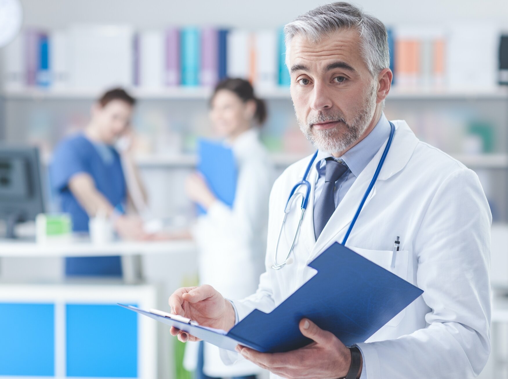 A male doctor with gray hair and a beard, wearing a white lab coat and stethoscope, holding a blue clipboard in a hospital or clinic with medical staff in the background.